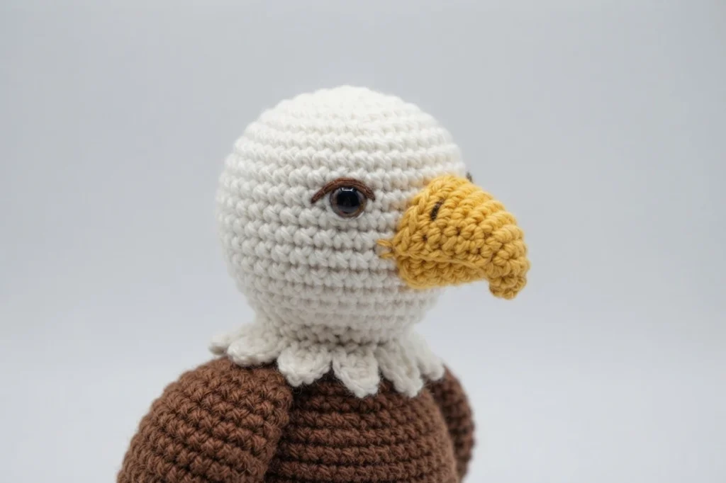 Close-up of a finished crochet eagle with a white head, a large yellow hooked beak, a brown body and white neck feathers on a plain light background