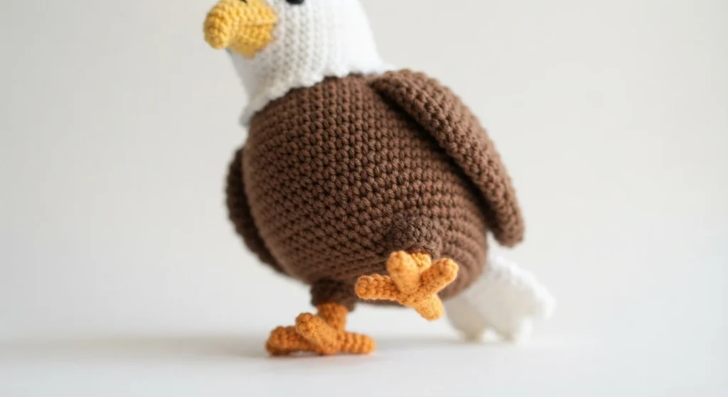 Close side view of a finished crochet eagle with a round brown body, brown wing, white head and orange feet