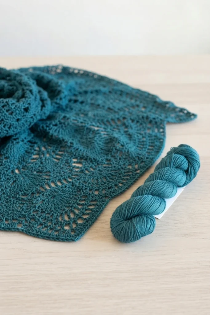 Teal crocheted lace shawl folded on a light wood surface beside a skein of matching teal yarn, showing open fan and shell motifs.