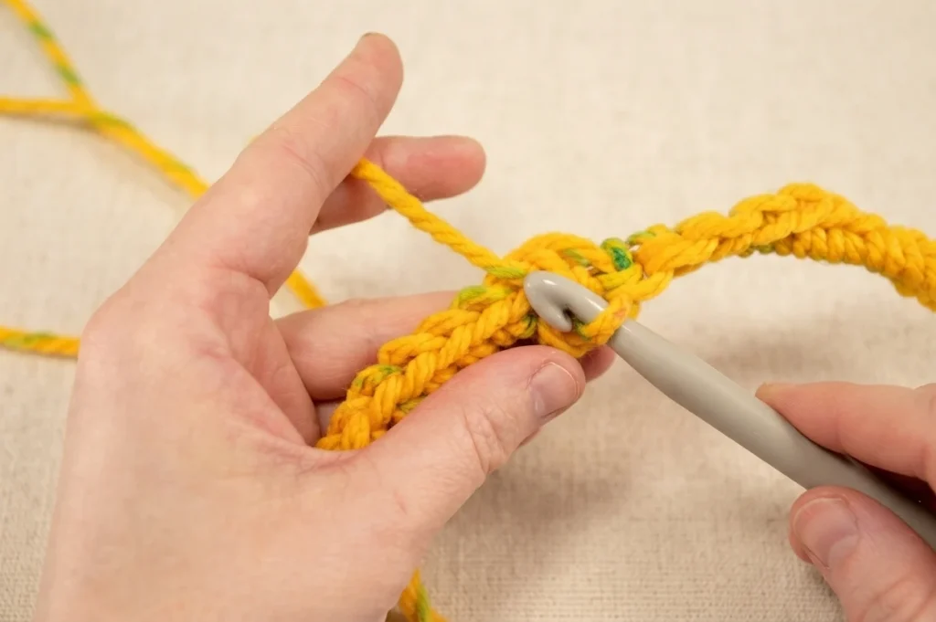 Close-up of hands crocheting with yellow yarn around a metal hoop using a gray crochet hook.