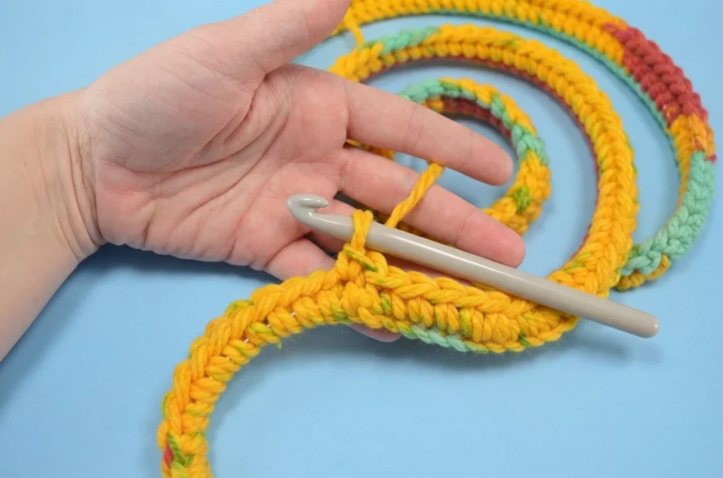 A hand holds a grey crochet hook and works yellow and green yarn into a spiral of single crochet stitches on a light blue background.