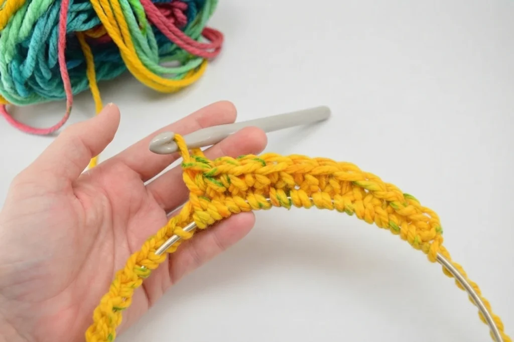 Hand holding a yellow crocheted strip worked around a metal hoop, with a crochet hook and variegated yarn in the background.