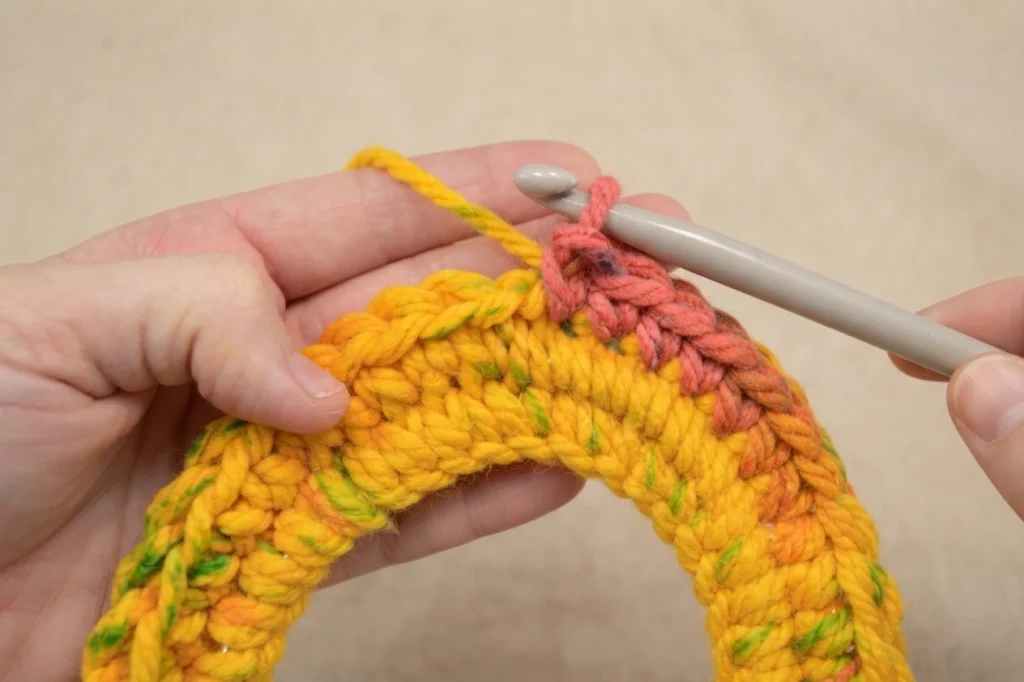 A hand working single crochet stitches in a spiral with a gray crochet hook into a colorful yellow, pink and green crocheted tunnel around a hoop