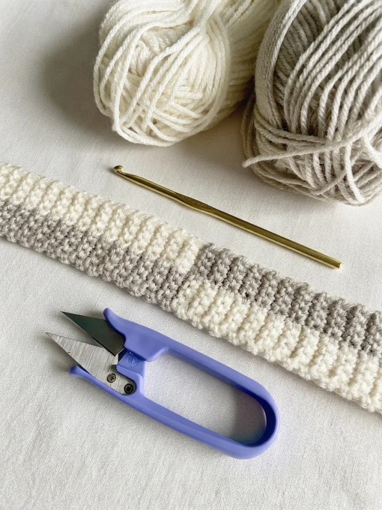 Crocheted bag strap in a cream and beige checker pattern lying diagonally beside cream and beige yarn balls, a gold crochet hook and blue yarn snips