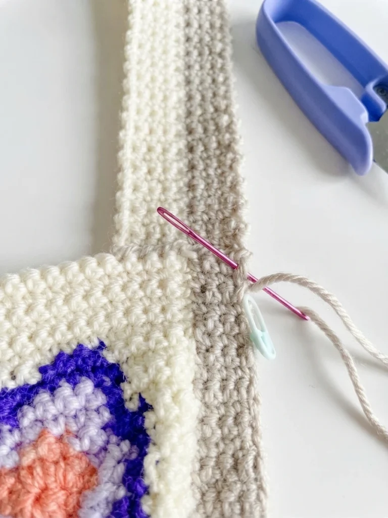 Close up of a two tone crochet strap being sewn to a butterfly bag panel with a yarn needle, a stitch marker and thread snips nearby