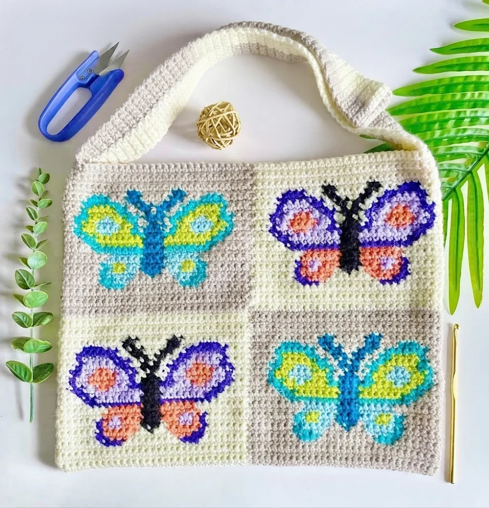 Finished crochet bag with four butterfly motifs in teal, yellow, purple and peach, a cream strap, scissors, a crochet hook and green leaves