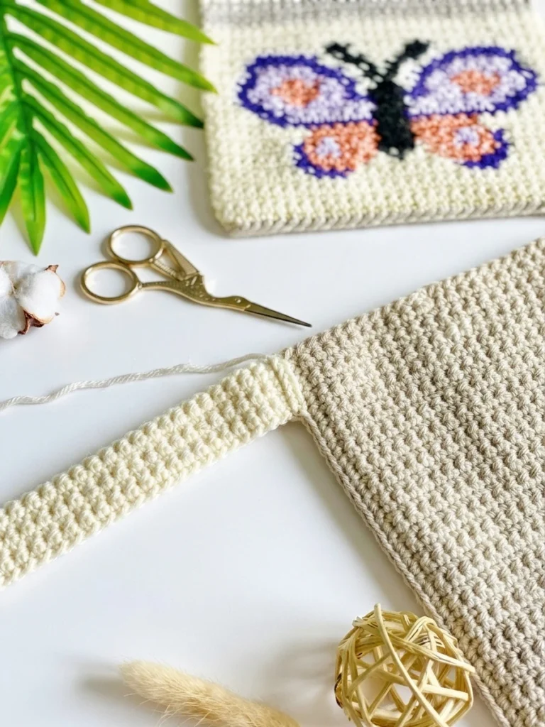 Close-up of a crocheted butterfly panel in purple, lavender and peach above a beige panel with a cream strap being attached, beside gold scissors and a green palm leaf