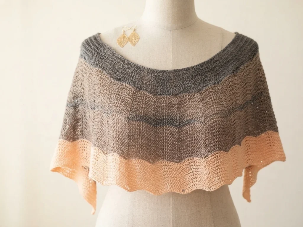 Châle au crochet gris, taupe et pêche drapé sur un mannequin, avec des motifs ajourés ondulés et une bordure festonnée.