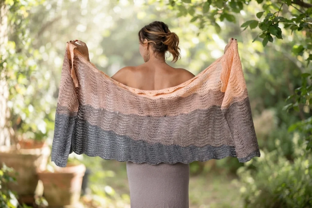 Une personne vue de dos tient un grand châle crocheté aux motifs ondulés, pêche en haut, taupe au centre et gris foncé au bord inférieur festonné.