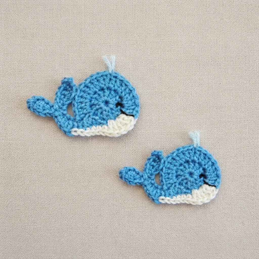 Two blue crochet whale appliques with white bellies and embroidered faces lying on beige fabric
