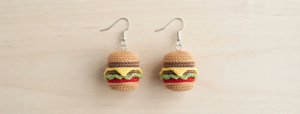 Pair of tiny crocheted burger earrings with tan buns, brown patty, yellow cheese, green lettuce and red tomato layers on silver fish hooks, hanging side by side against a light wood background