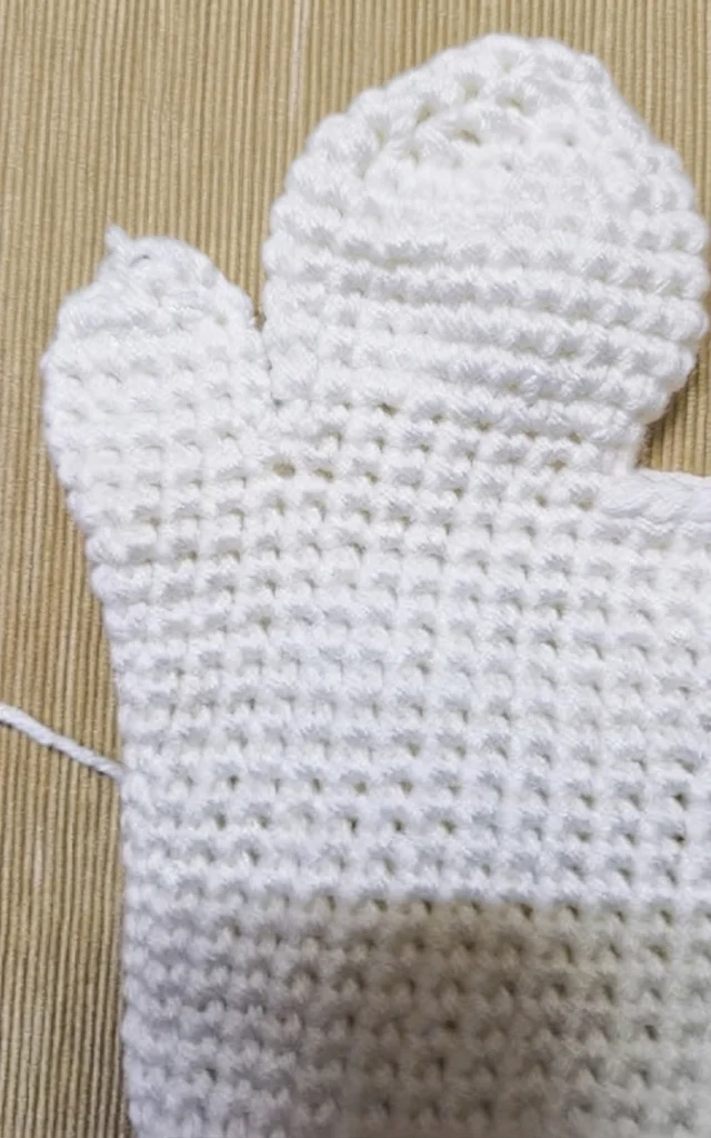 White crochet ghost hand puppet body in progress on a beige textured surface, showing a rounded head section, a thumb opening on the left, a small yarn tail at the side and no face yet
