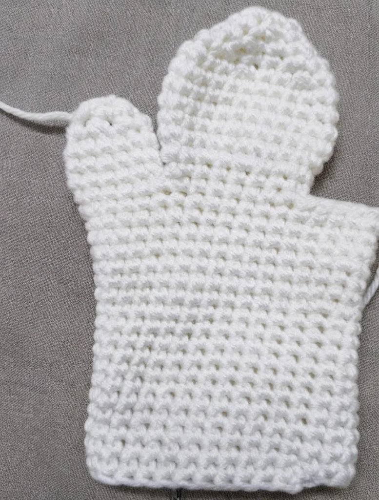 White crochet ghost hand puppet body in progress, with a rounded head, a thumb opening and loose yarn tails, laid flat on a gray fabric background.