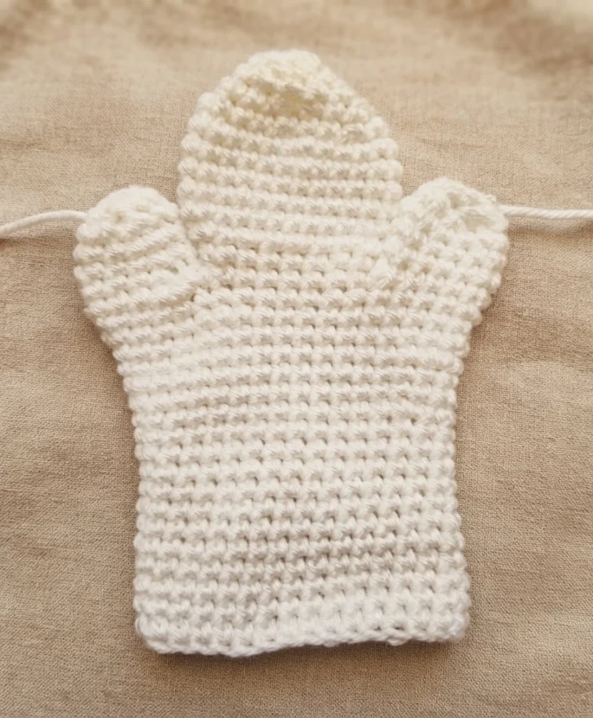 White crocheted hand puppet body in progress, shaped like a ghost with a rounded head and two side openings, loose yarn tails at both sides, lying flat on a beige textured fabric background