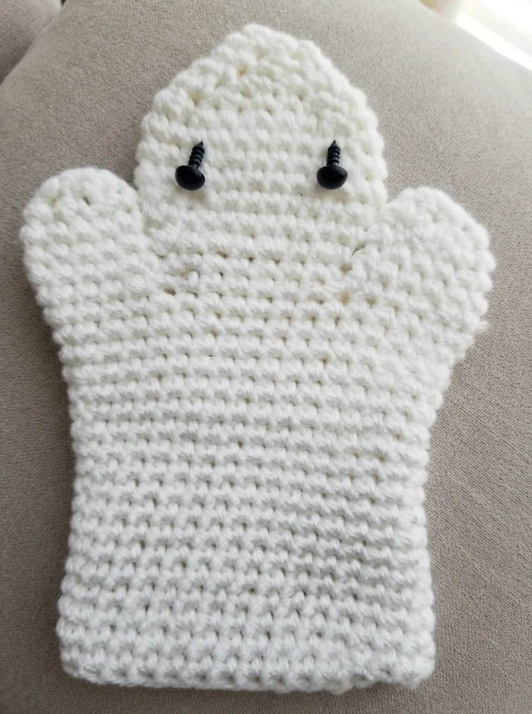 White crocheted ghost hand puppet with two black screw eyes, worked in single crochet with a rounded head, two side arms and a flat rectangular body, laid on a beige fabric surface.