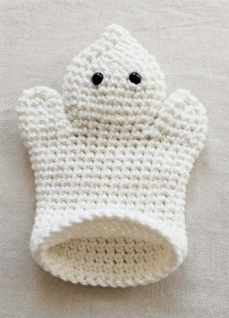 White crocheted ghost hand puppet with two black bead eyes, lying flat with the open bottom edge showing the hollow inside, on a light beige fabric background.