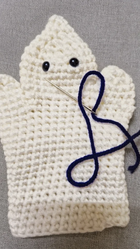 Crocheted white ghost hand puppet with two black bead eyes, a tapestry needle threaded with navy yarn resting against its body while the mouth is being embroidered, shown flat on a gray textured background.