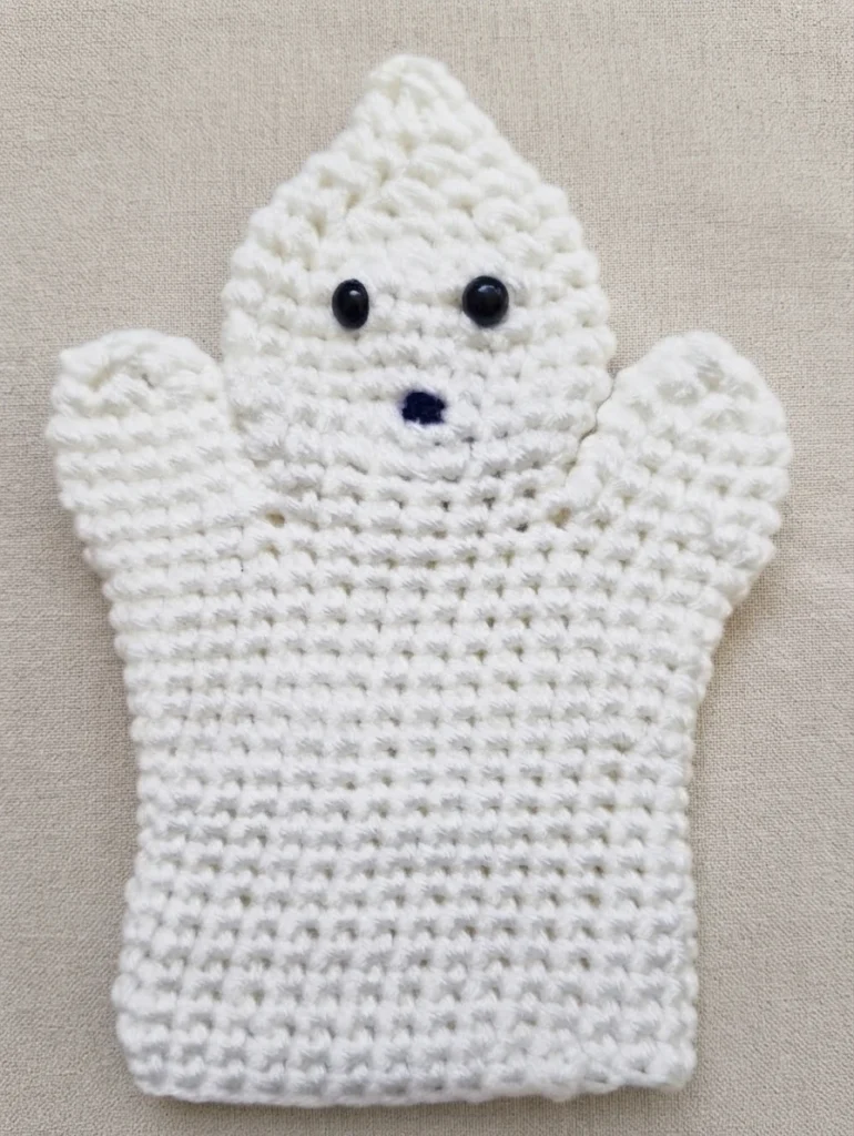 White crochet ghost hand puppet lying flat on a light beige fabric, with two black bead eyes, a small dark stitched mouth and two raised side arms.