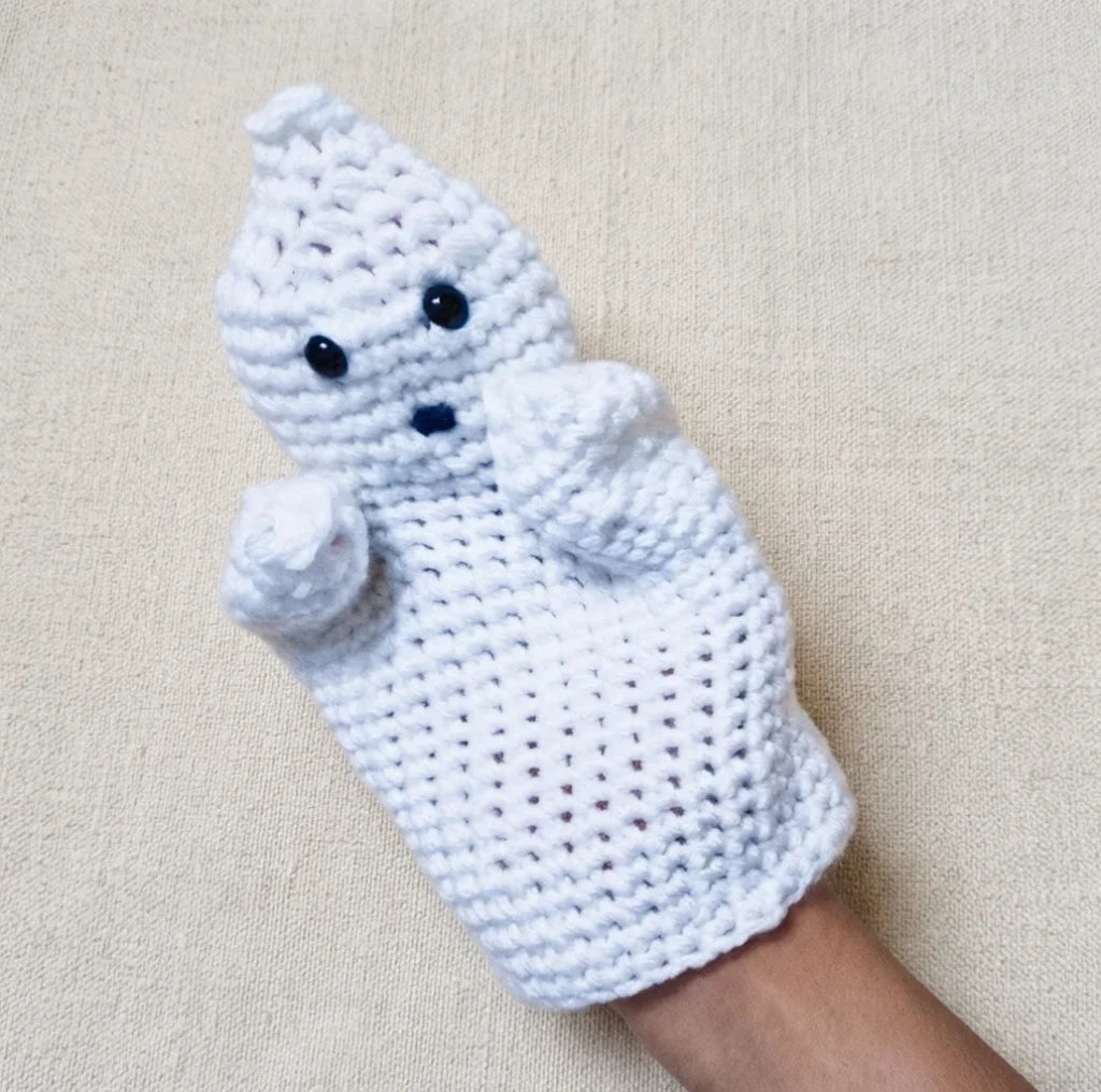 White crocheted ghost hand puppet worn on a hand, with two black bead eyes and a small dark mouth, shown against a plain cream fabric background