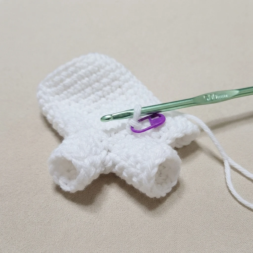 White crochet ghost hand puppet in progress on a beige background, with the thumb and pinky finger openings already formed, a green crochet hook inserted in the working loop and a purple stitch marker holding a stitch.