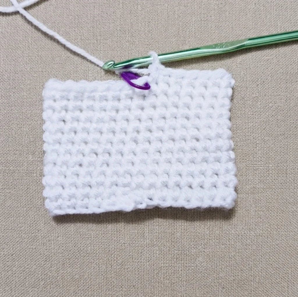 White single crochet swatch in progress on a beige textured fabric, with a green crochet hook and a purple stitch marker at the top working edge