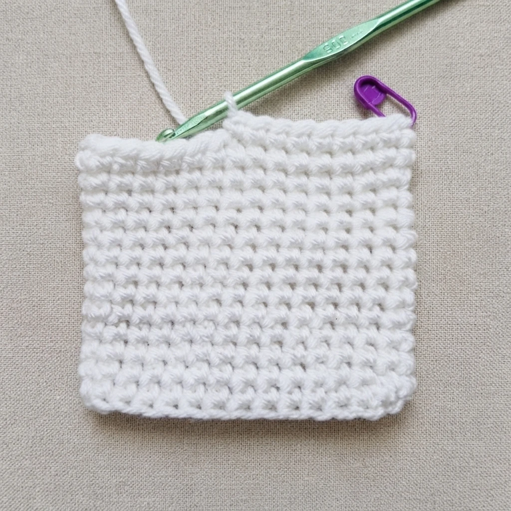 White crocheted square piece in progress on a green crochet hook with a purple stitch marker, resting on a beige woven fabric.