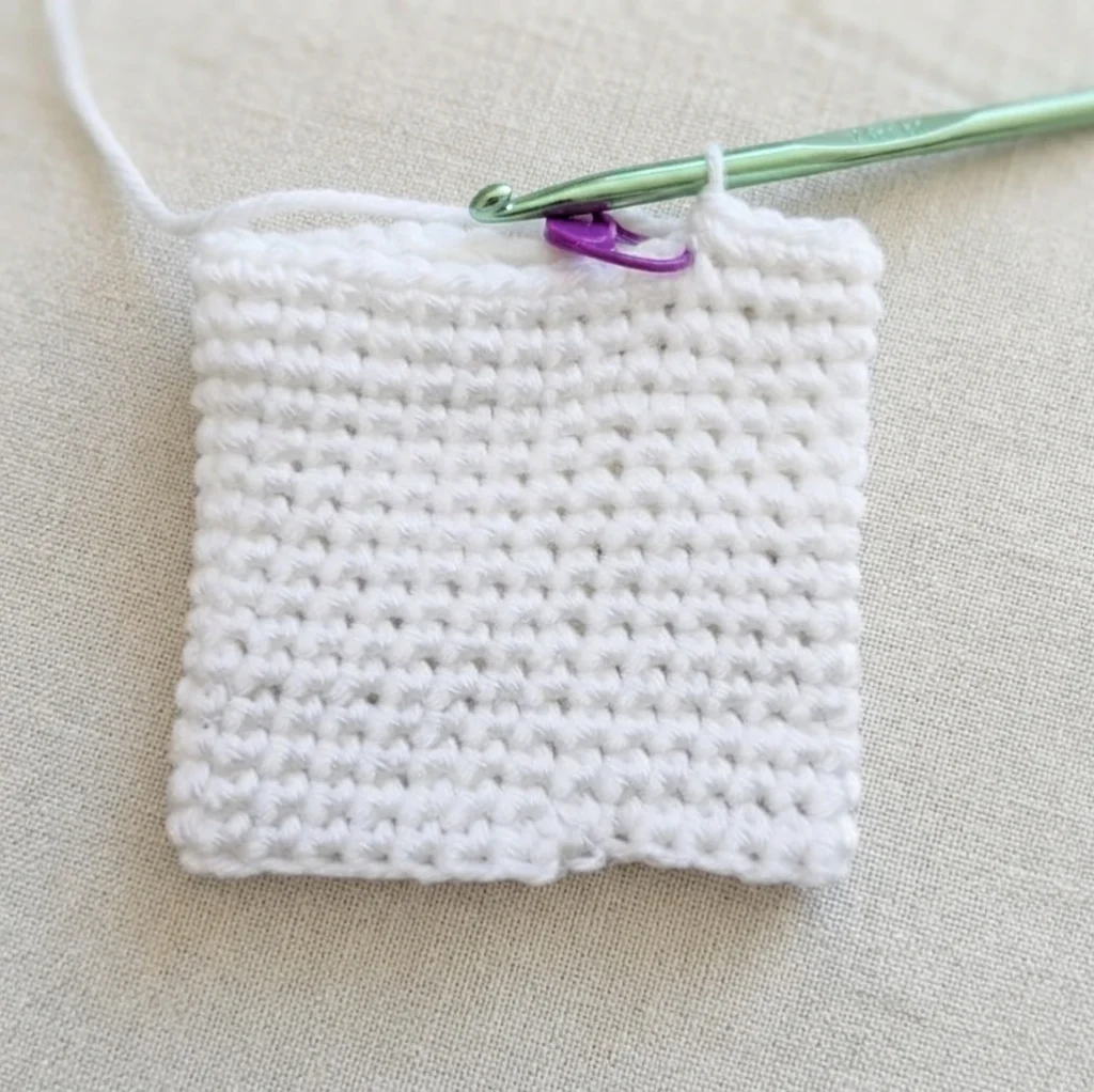 Carré de crochet blanc en cours de réalisation, crochet métallique vert et marqueur de maille violet posés dessus, sur une toile beige clair