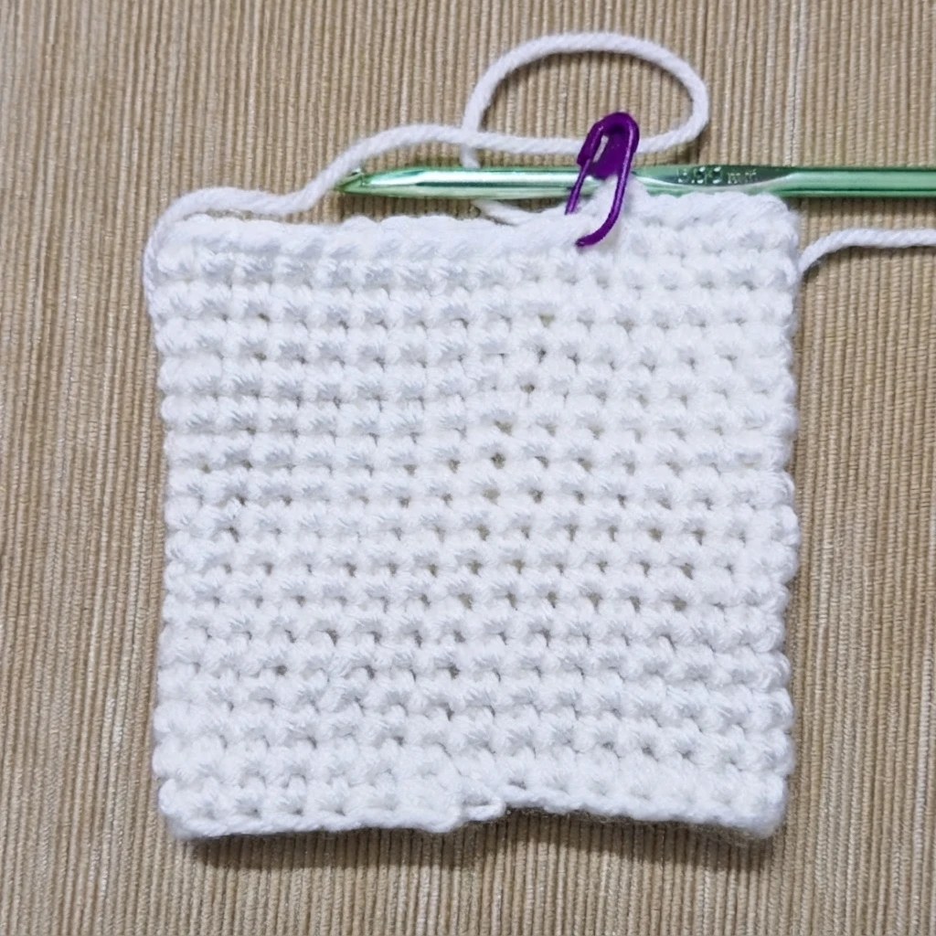 White single crochet swatch in progress on a tan textured surface, with a green crochet hook along the top edge and a purple stitch marker holding the working loop.