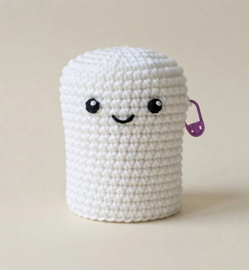 White crocheted piece in progress shaped as a rounded cylinder, with two black embroidered eyes, a small black embroidered smile and a purple stitch marker clipped to the side, on a plain beige background.