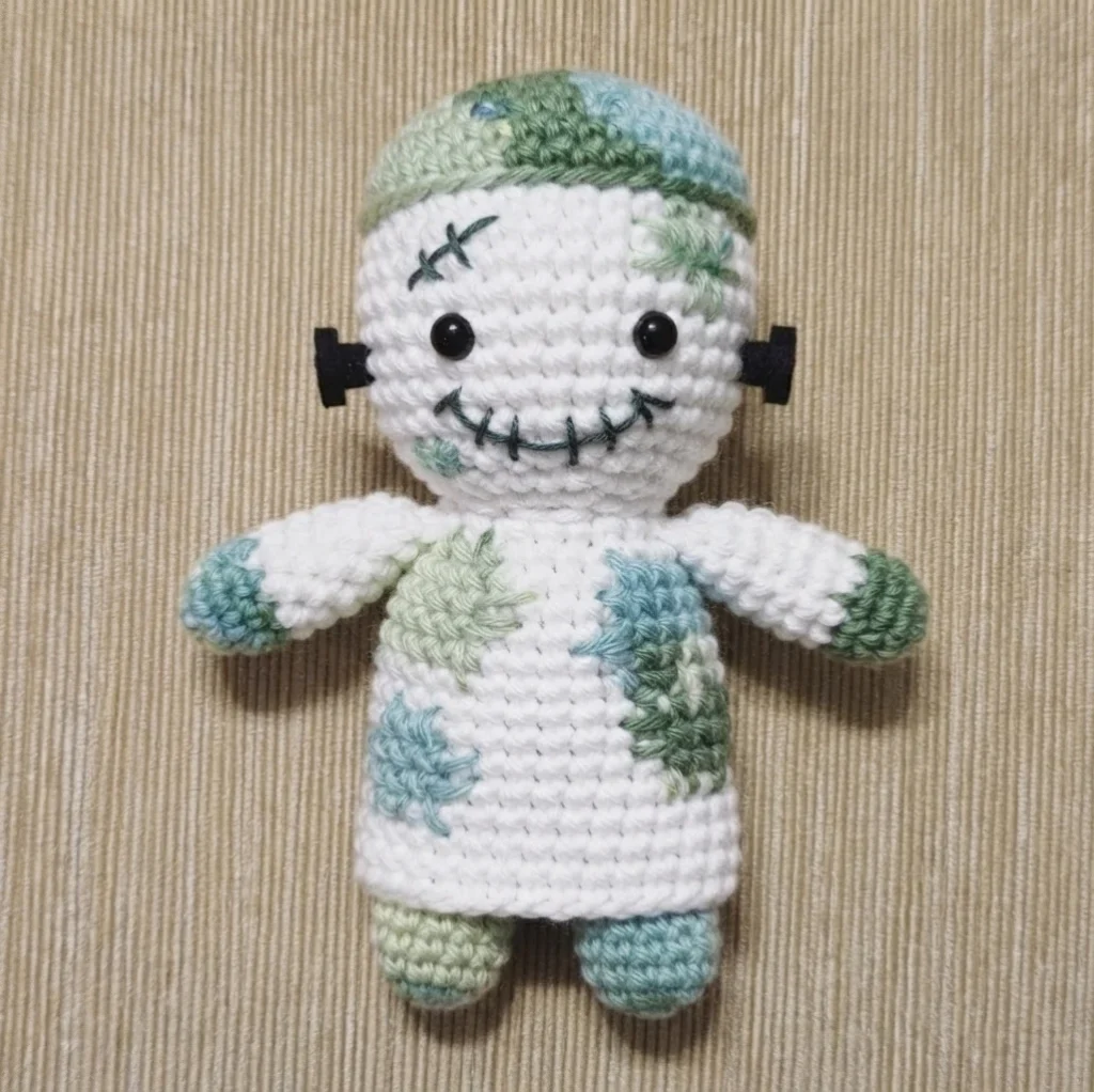 Crocheted white doll with green and mint yarn accents, round black bead eyes, a stitched dark mouth and cross stitch above one eye, black bolt shapes on both sides of the head, small arms and legs, standing on a beige striped surface.