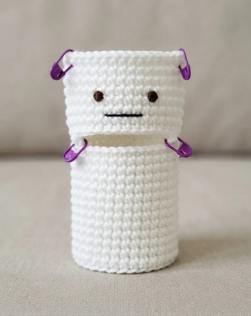 White crochet ghost hand puppet in progress standing upright, with two small brown stitched eyes and a straight black stitched mouth, four purple stitch markers holding the top and middle openings apart, on a plain beige background.