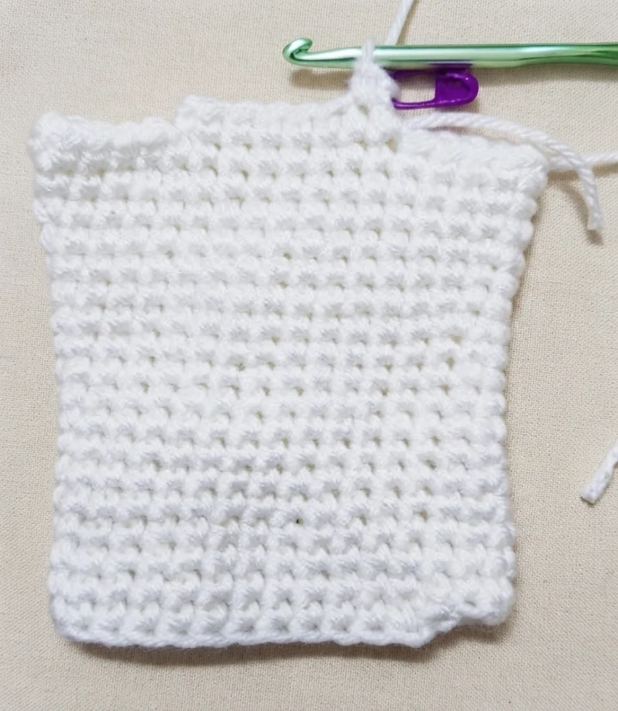 White crocheted rectangle in progress on a plain tan fabric surface, with a green crochet hook and a purple stitch marker resting at the top edge, loose yarn tails on the right side and slightly uneven top and bottom edges.