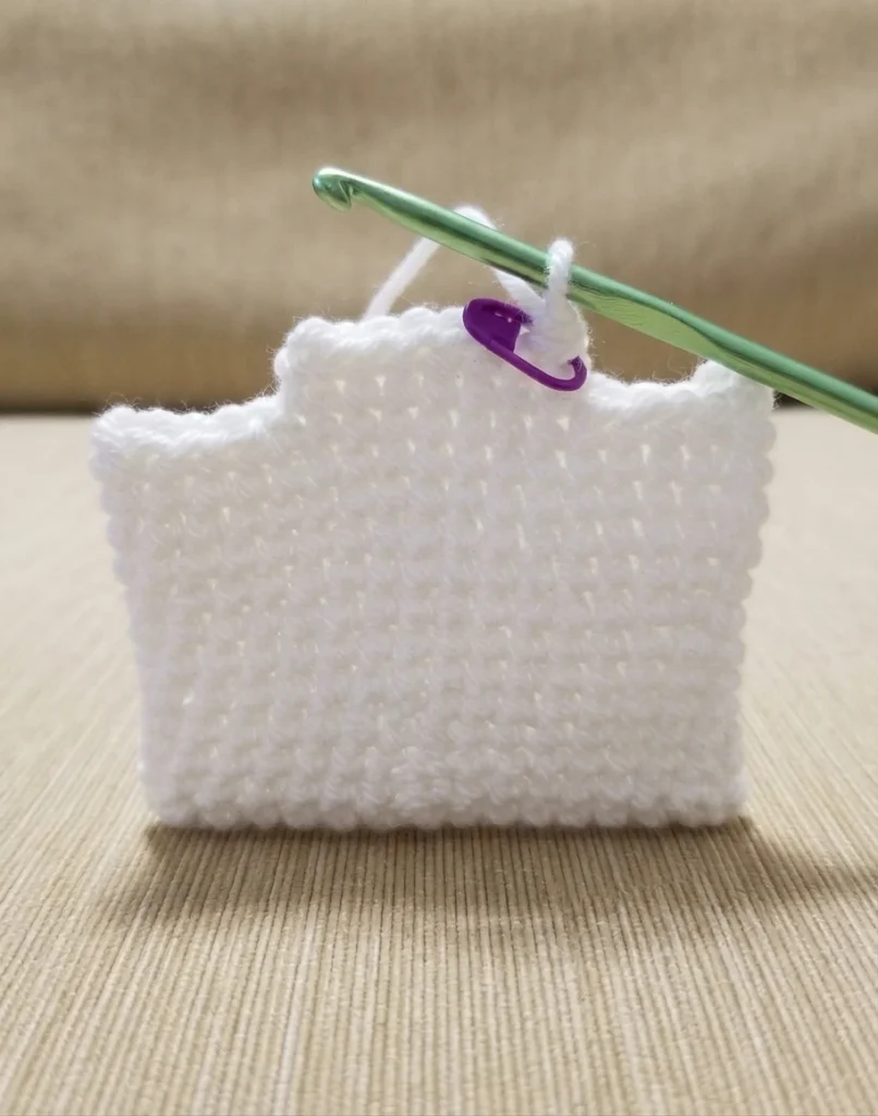 White crochet work in progress on a green hook with a purple stitch marker, resting on a beige textured surface