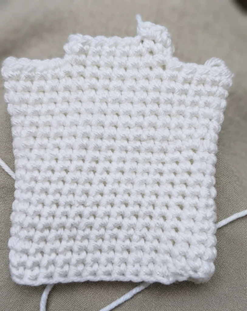 White crocheted ghost hand puppet body in progress, a flat rectangular panel of single crochet with two small bumps along the top edge and loose yarn tails at the sides, lying on a beige fabric background.