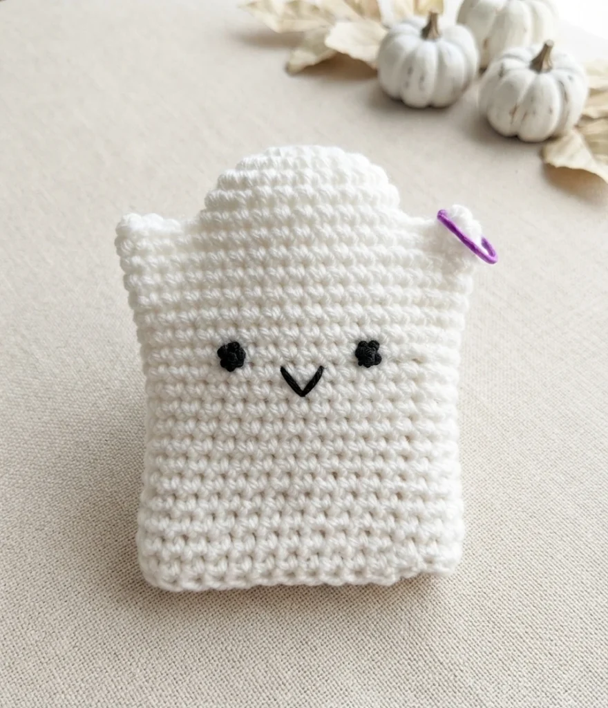 White crochet ghost hand puppet with black embroidered dot eyes and a small V-shaped smile, a purple stitch marker on the right edge, white pumpkins and dried leaves blurred in the background on a light fabric surface.