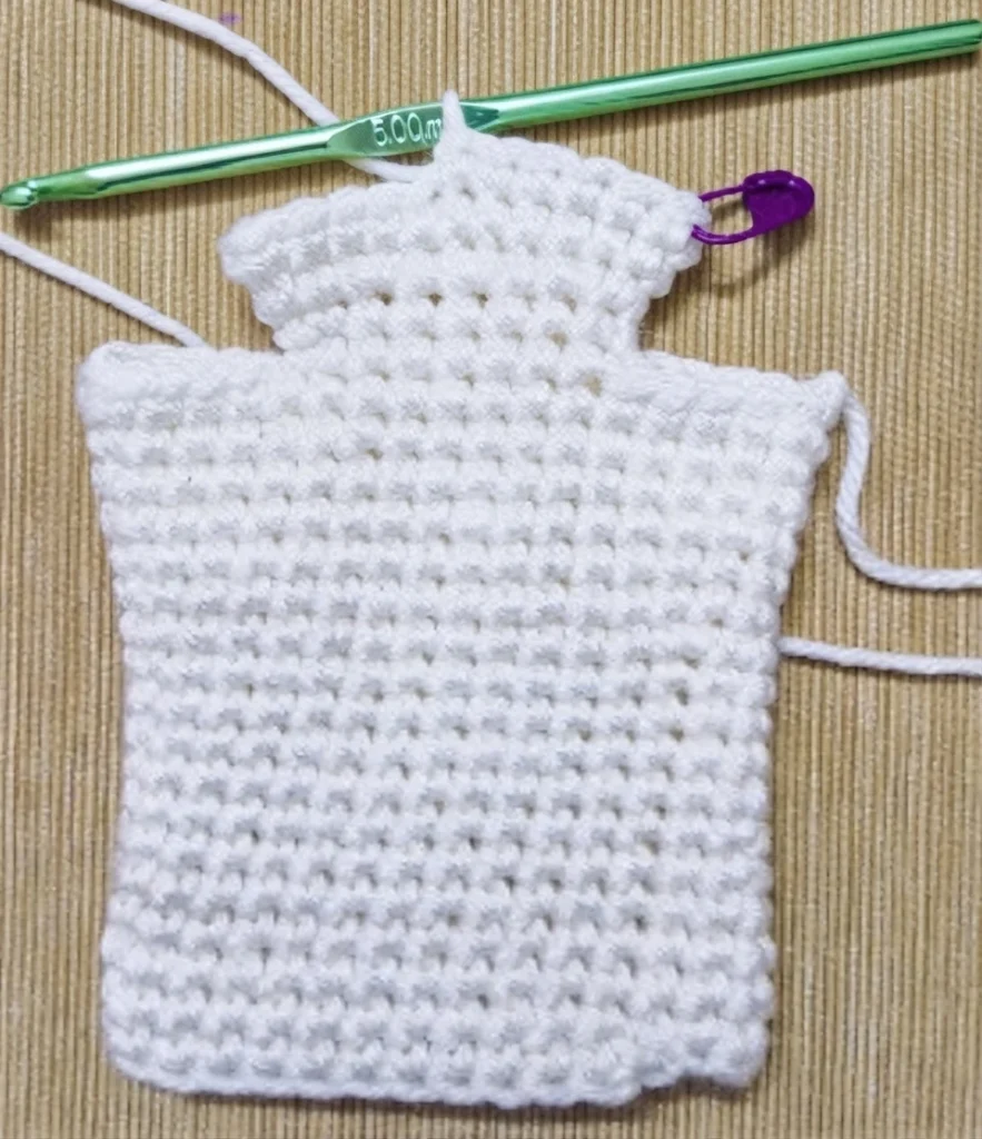 White crochet ghost puppet body in progress on a ribbed brown mat, with a green crochet hook resting above it, a purple stitch marker holding the working loop and loose yarn tails at the sides.