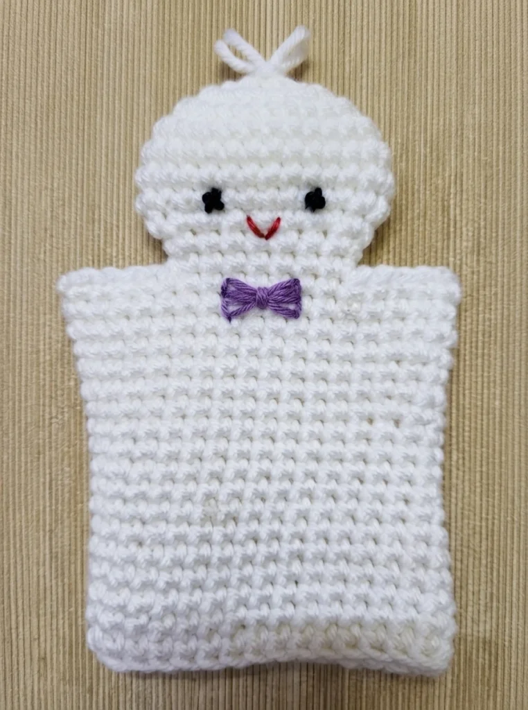 White crochet ghost hand puppet with black bead eyes, a small red stitched smile and a purple yarn bow, with a white hanging loop at the top of the head, lying flat on a tan ribbed surface.