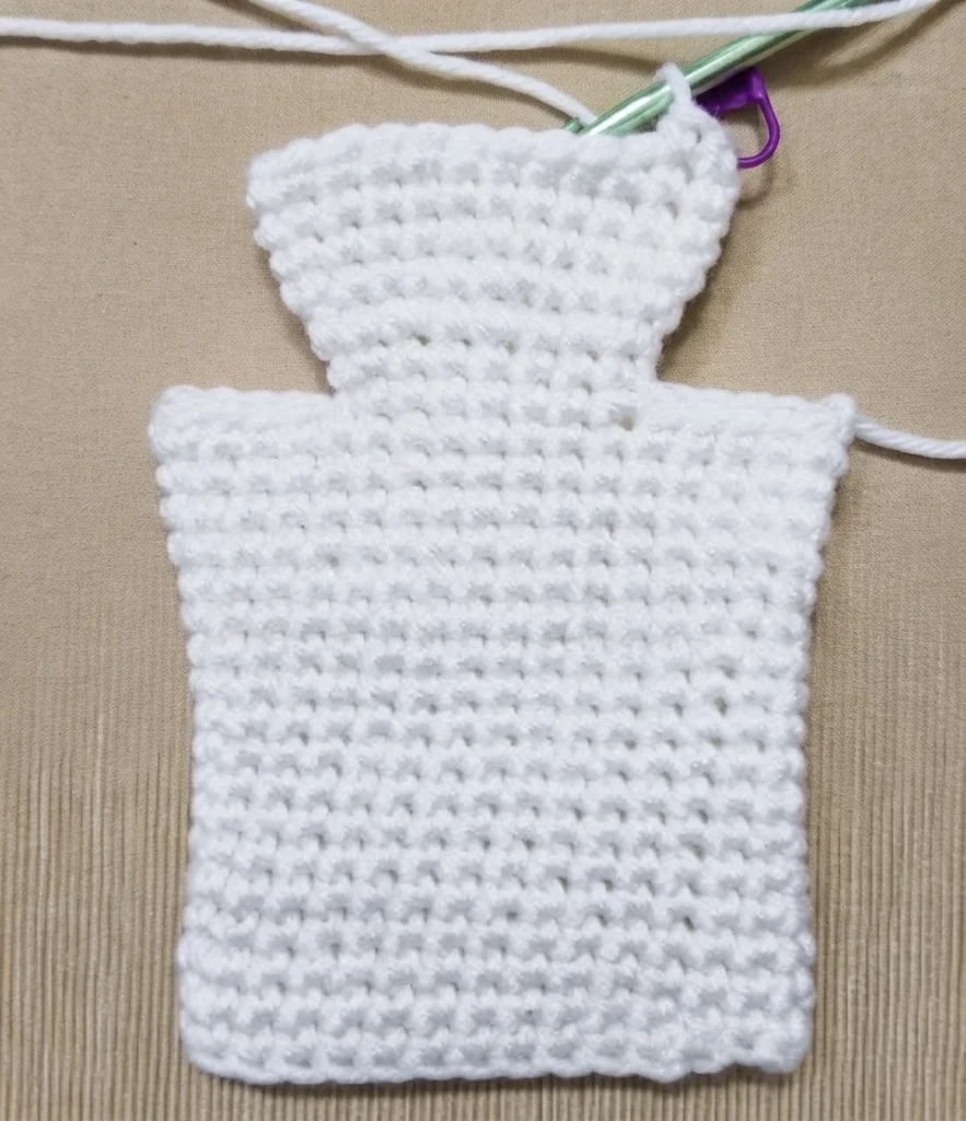 White crocheted ghost hand puppet in progress on a beige textured surface, showing a shaped head and neck above a rectangular body, with a green-handled crochet hook, working yarn and a purple stitch marker at the top.