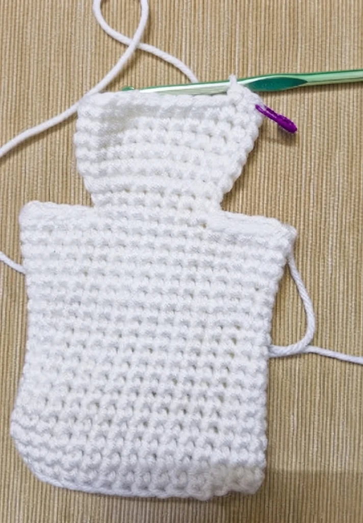 White crocheted ghost hand puppet in progress on a beige textured mat, with a green crochet hook resting across the top and a purple stitch marker, showing the head and body shape before the eyes and mouth are added.