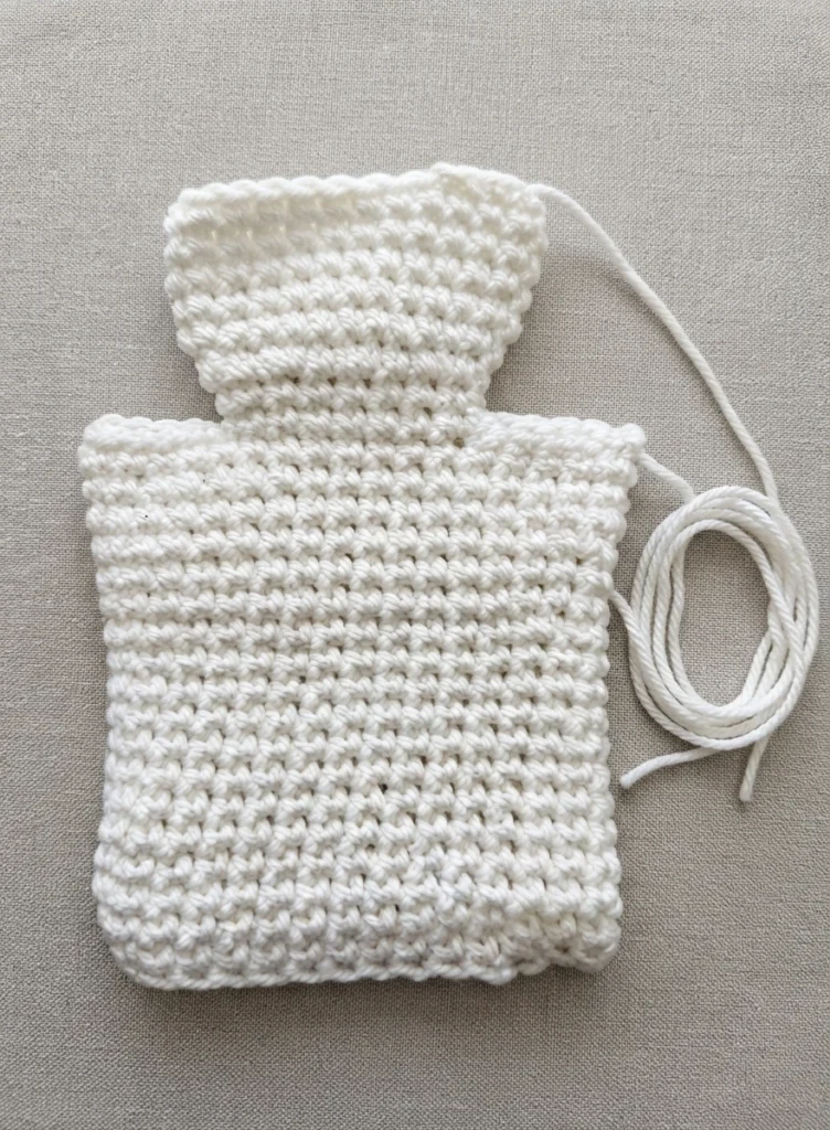 White crocheted ghost hand puppet body in progress, with a rounded head and rectangular single crochet body, a loose strand of white yarn coiled at the right side, resting on a light grey fabric background.