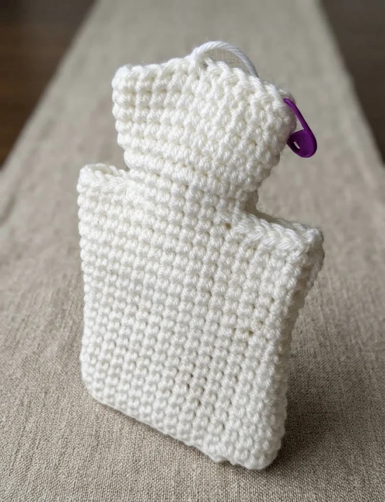 White crocheted ghost hand puppet shape in progress on a beige fabric surface, with a purple stitch marker at the neck and a short yarn tail at the top.