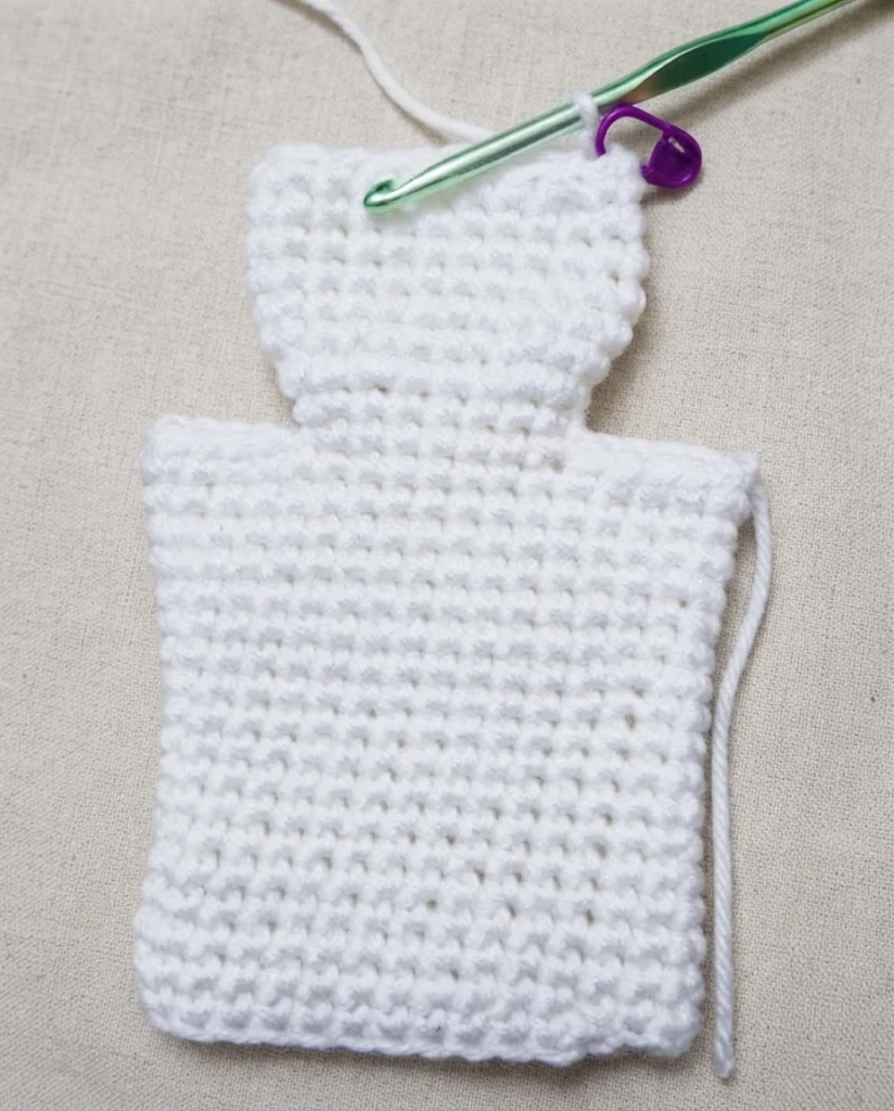 White crocheted ghost hand puppet in progress, laid flat with a green crochet hook and a purple stitch marker at the top opening, on a light beige fabric background.
