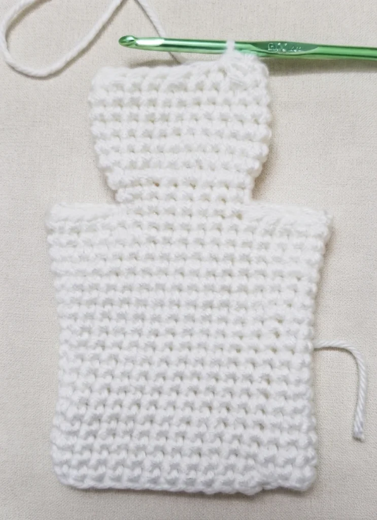 White crocheted ghost puppet body in progress on a light cream fabric background, with a green metal crochet hook resting at the top, a loose strand of white yarn above it and a yarn tail hanging from the right side.