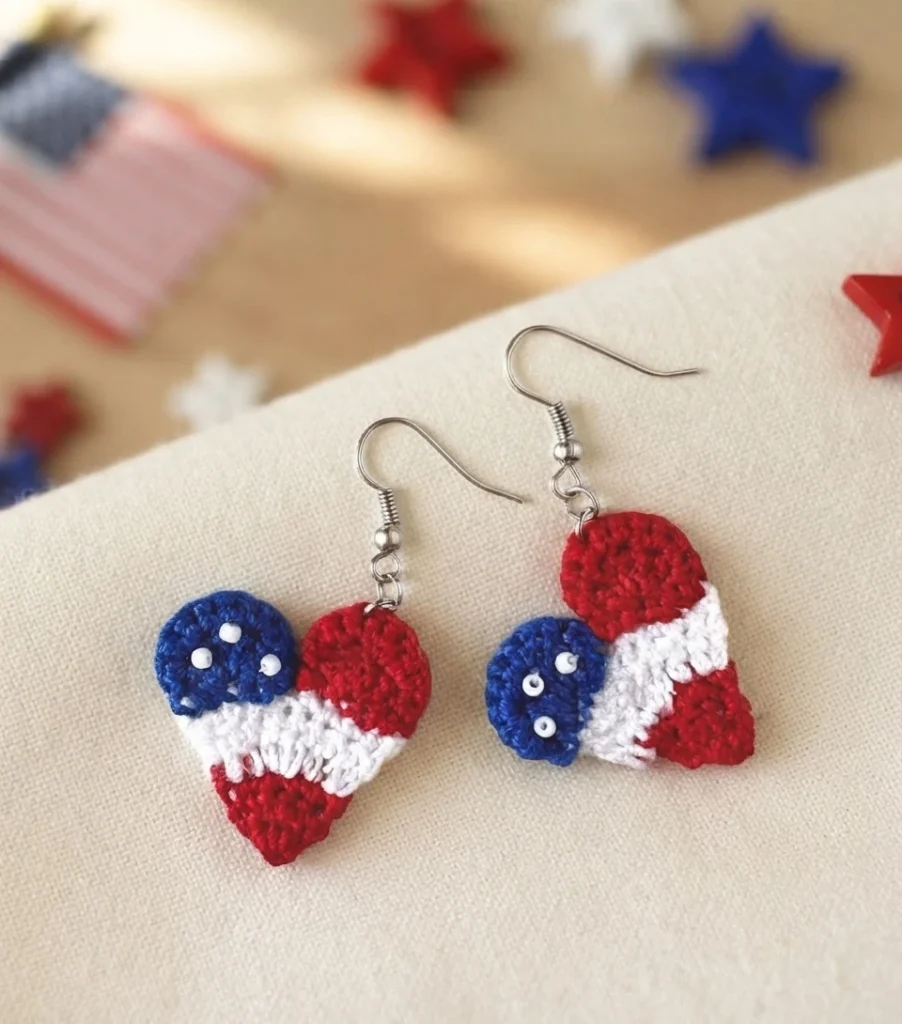 Pair of crochet heart earrings in red, white and blue with white beads on the blue section, on a light fabric background with blurred stars