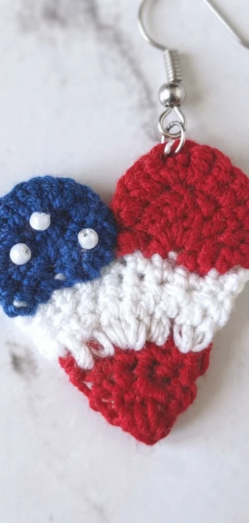 Close-up of a single crochet heart earring in blue, white and red with white beads on the blue section and a silver hook