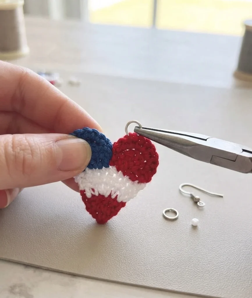 A hand holding a red, white and blue crochet heart earring while pliers attach a jump ring, with an earring hook and spare jump ring nearby