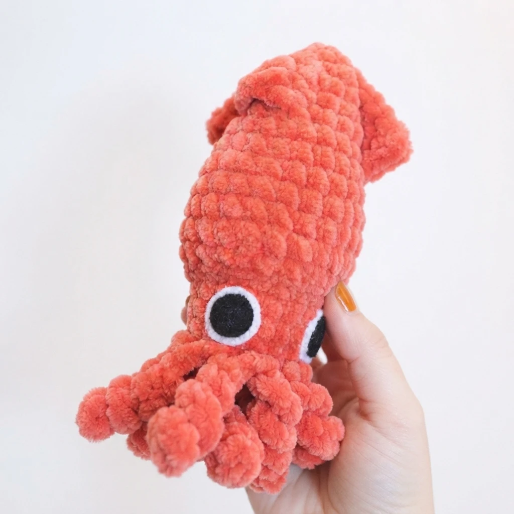 Hand holding a coral crochet squid with black and white felt eyes and small folded fins against a white background