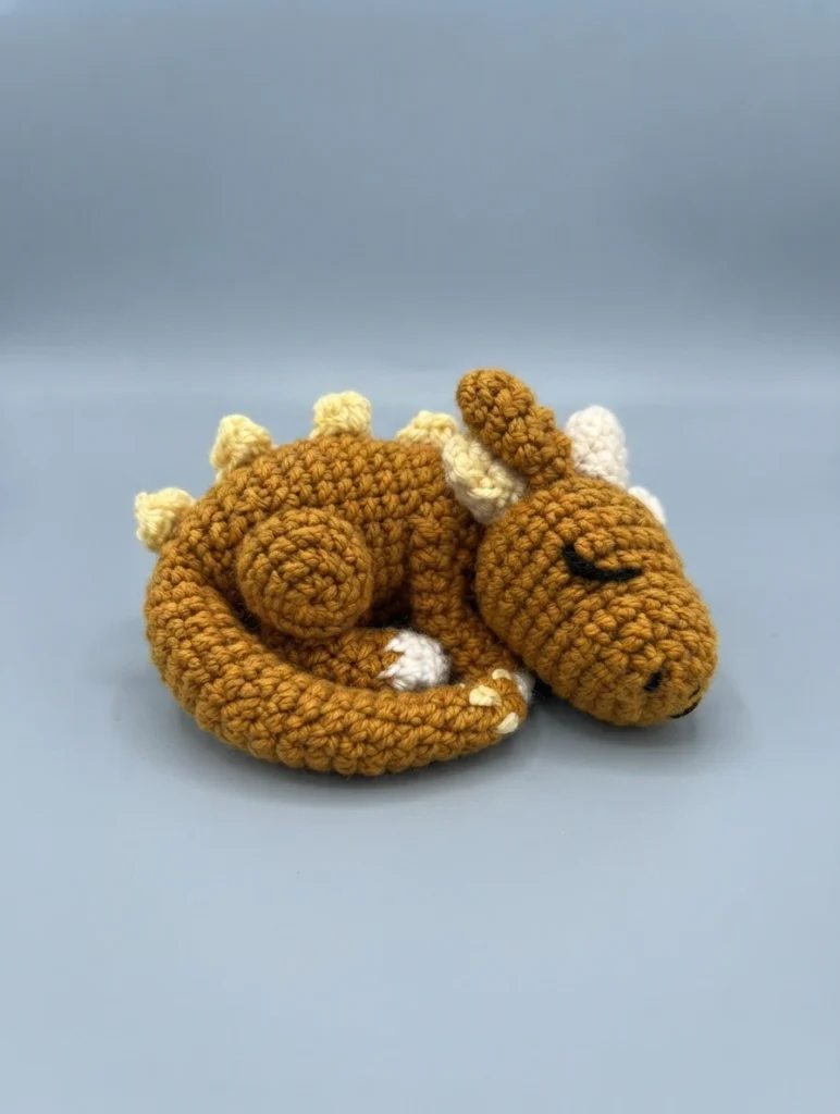 A mustard brown crochet dragon curled up asleep with cream horns, pale yellow back spikes and a closed embroidered eye on a plain light blue grey background