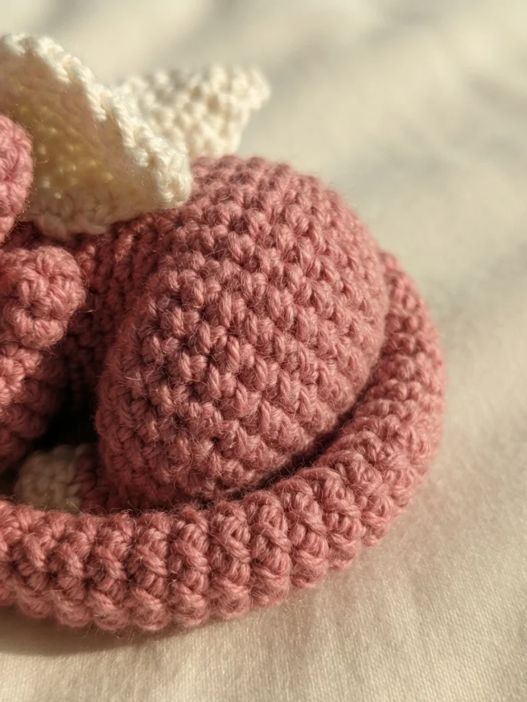 Close-up of a dusty pink crocheted dragon curled up with a cream wing and its tail wrapped around its body on a light fabric background