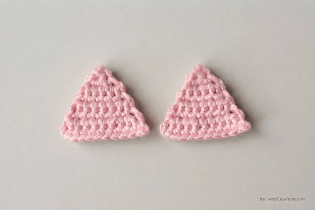 Two small pink crocheted triangular inner ear pieces worked in rows, placed side by side on a plain light gray surface.